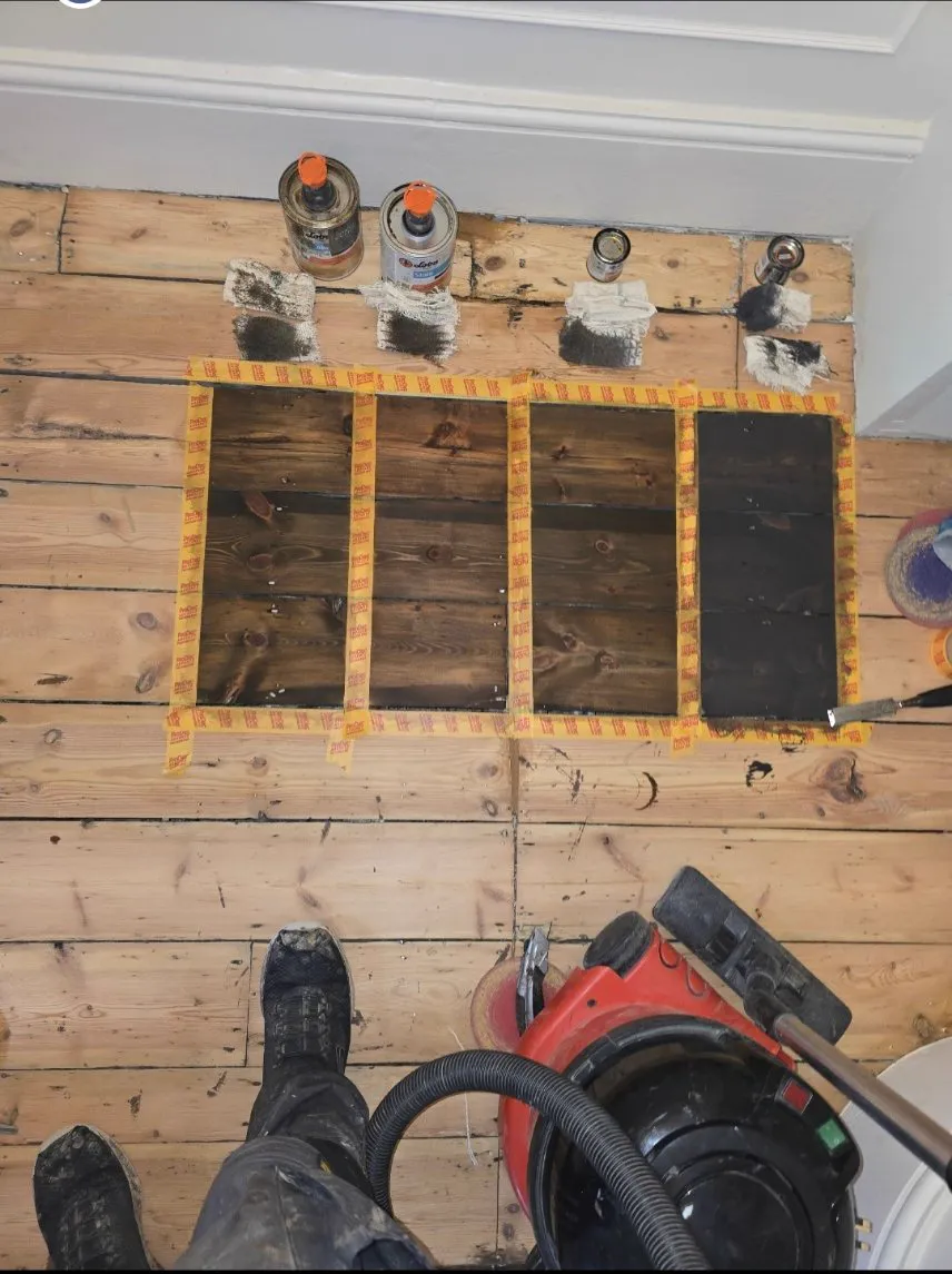 Stain patch test on original pine floorboards: four shades of brown taped off side by side with LOBA tins and sanding kit